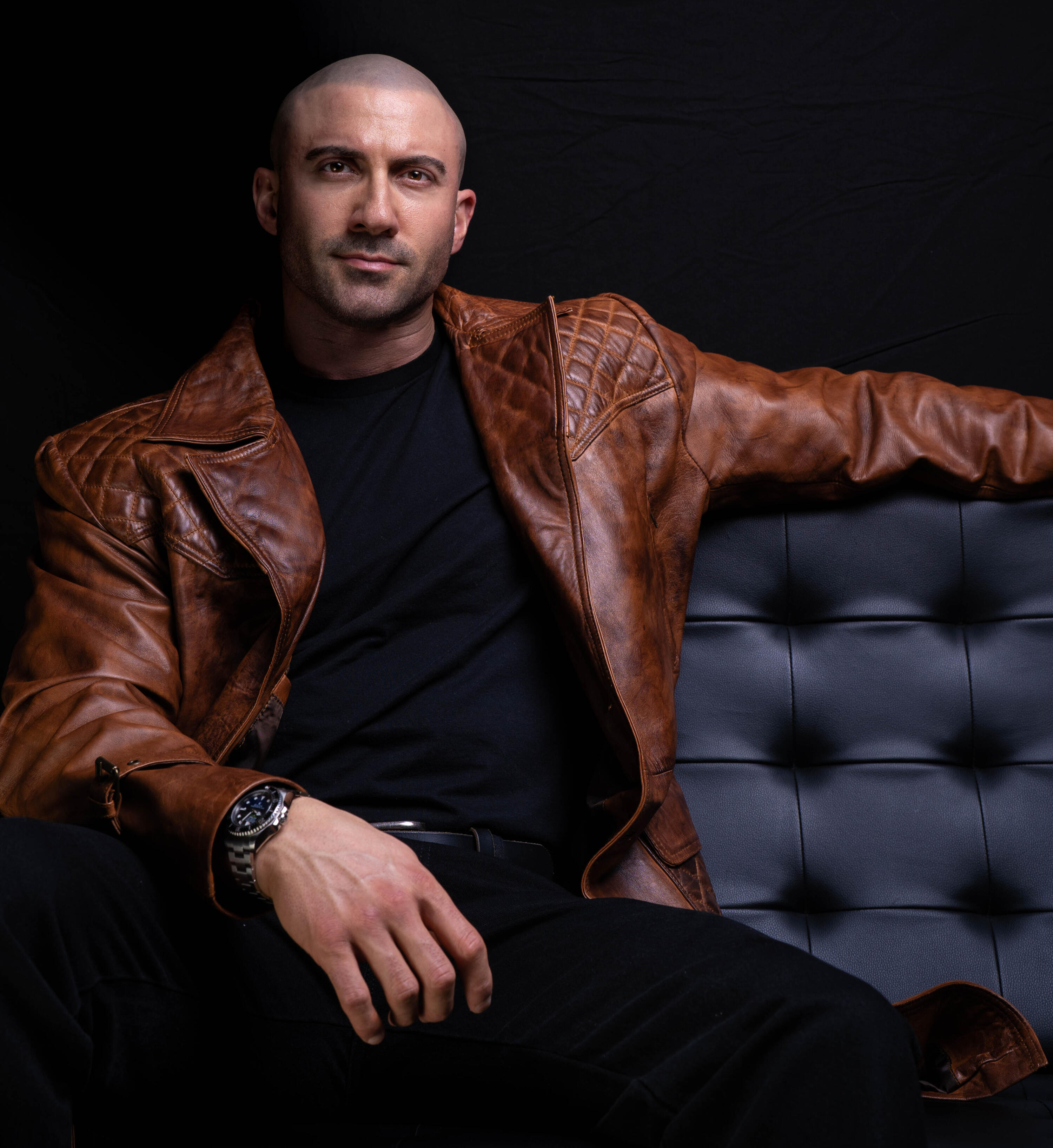 Author Thomas Rooney (Tom Rooney), sitting in a black Barcelona chair wearing long brown leather blazer, promoting Reflections of a Male Revue Headliner: How to Win at Life and Love